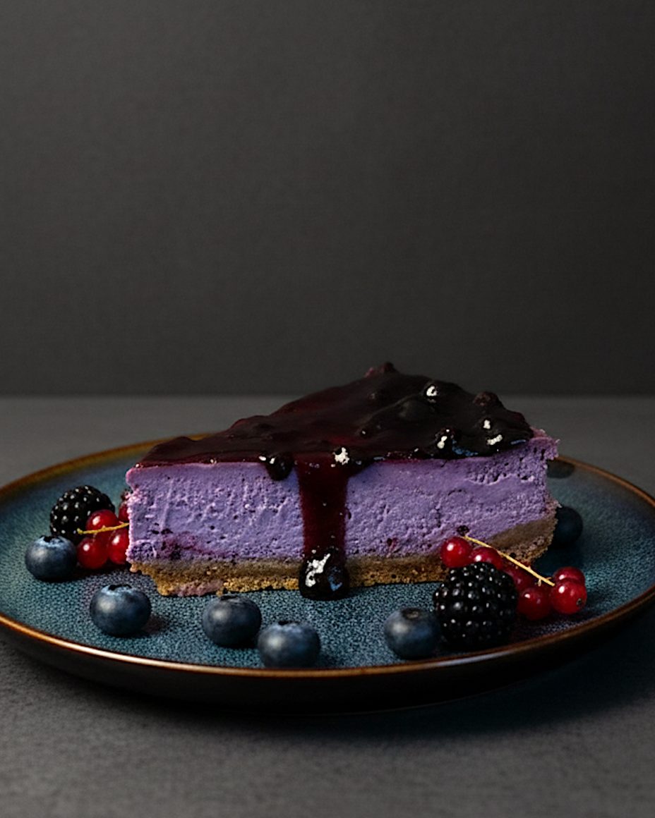 Une part de cheesecake Forestberry nappée de compote de fruits des bois sur une assiette bleu foncé, avec des myrtilles, des mûres et des groseilles