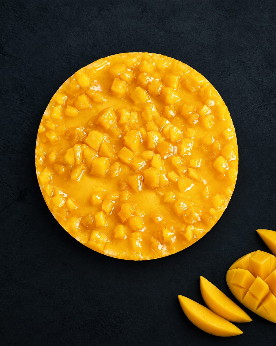 Cheesecake Mango New York entier vu de dessus, couvert de dés de mangue, avec une mangue coupée à côté