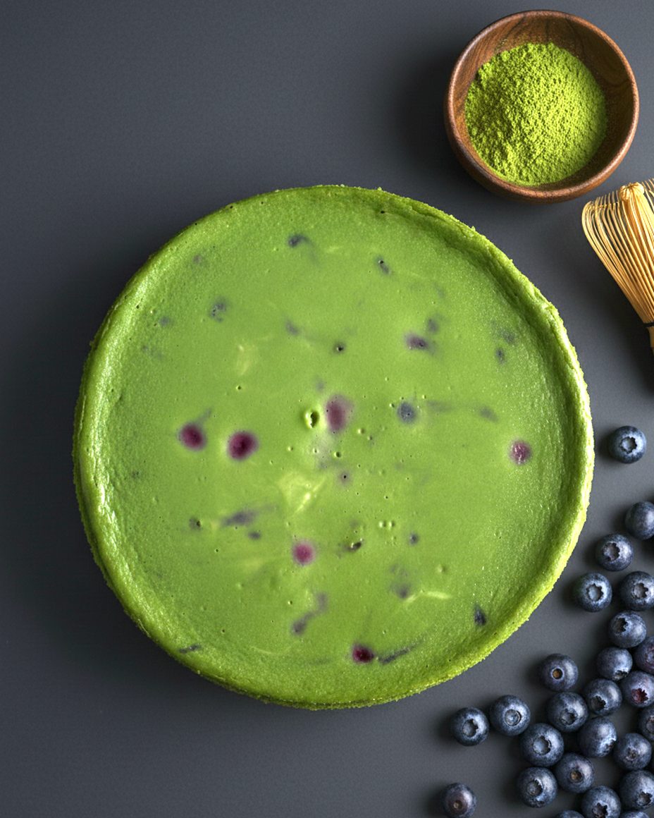 Cheesecake Matcha Ceremony entier vu de dessus, avec un bol de matcha en poudre, un fouet en bambou et des myrtilles