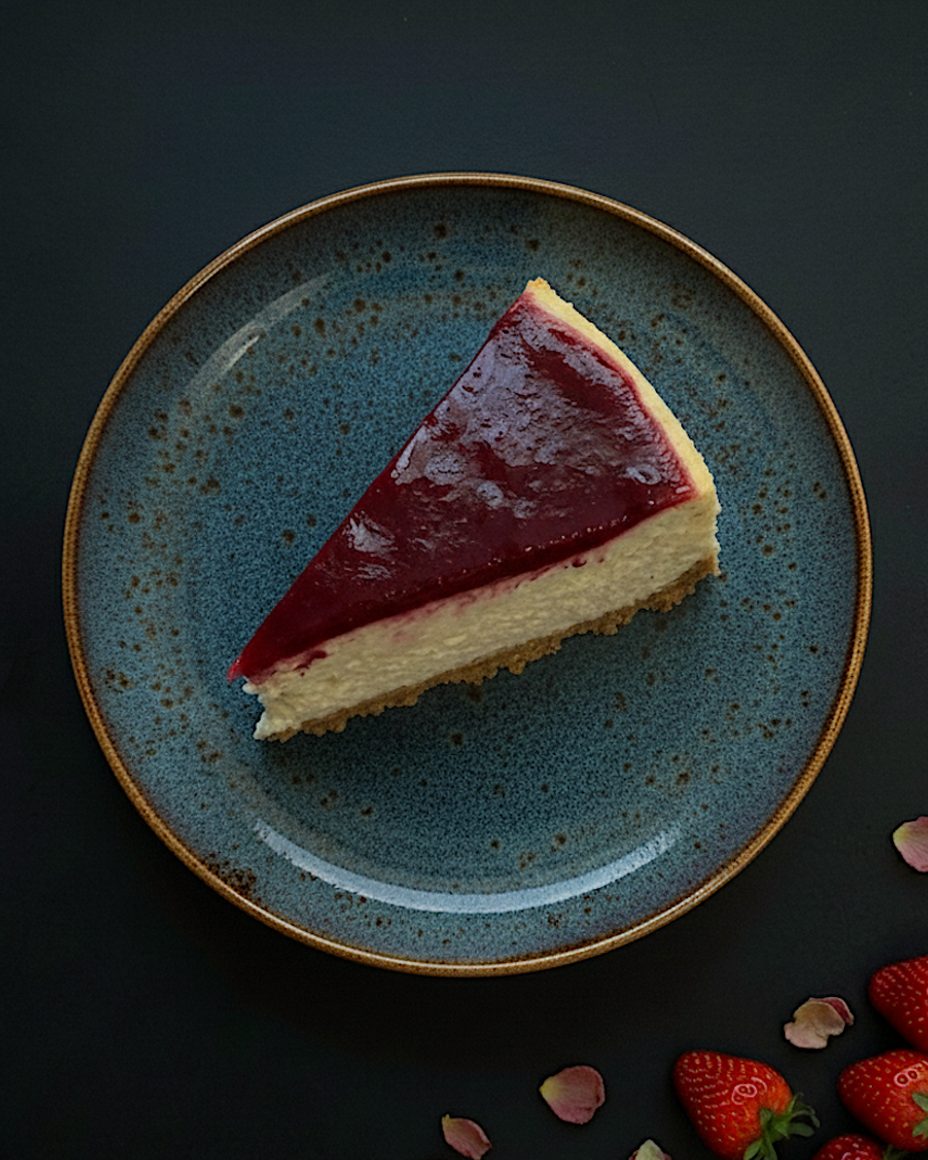 A slice of Strawberry New York cheesecake on a speckled blue plate, with fresh strawberries and rose petals