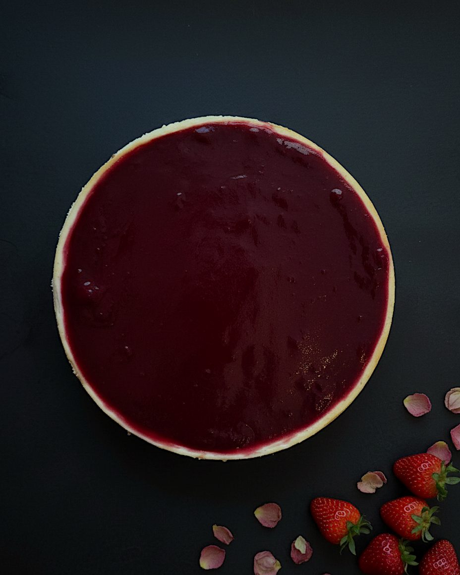 Strawberry New York cheesecake, whole cake seen from above, covered in strawberry compote, with fresh strawberries and rose petals